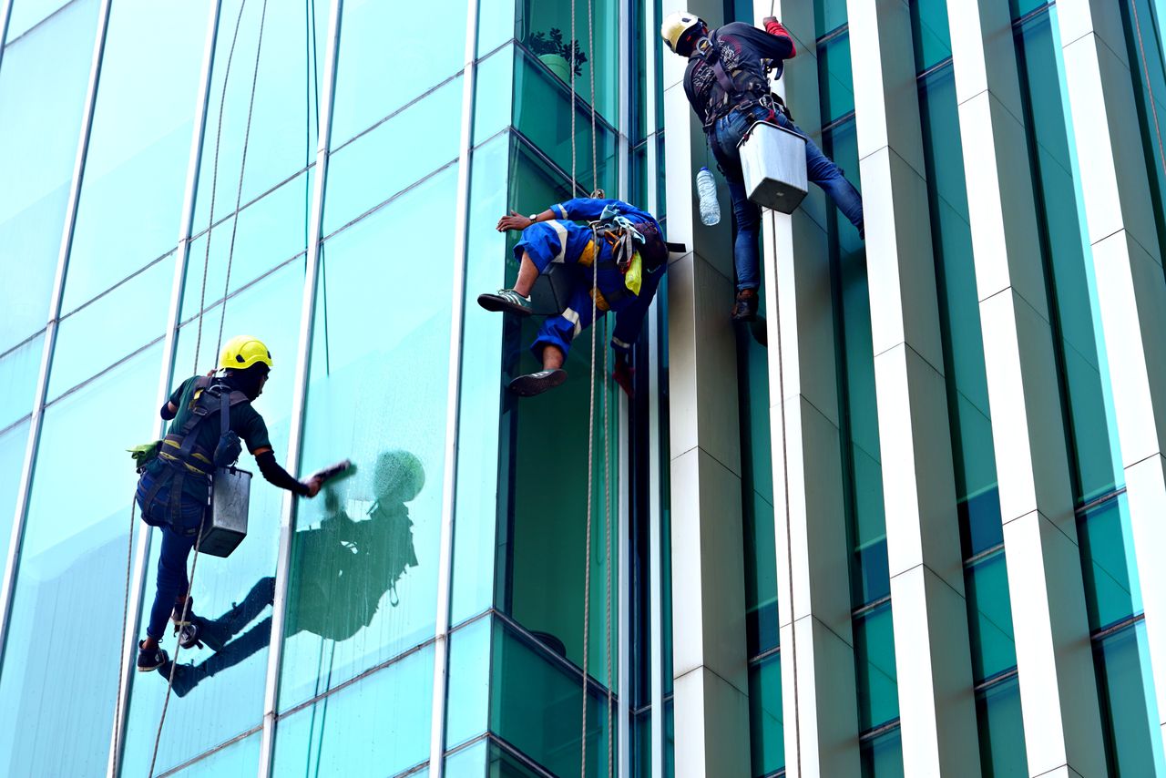 Trois laveurs de vitres, suspendus à des cordes, nettoient en journée la façade vitrée d'un immeuble moderne.